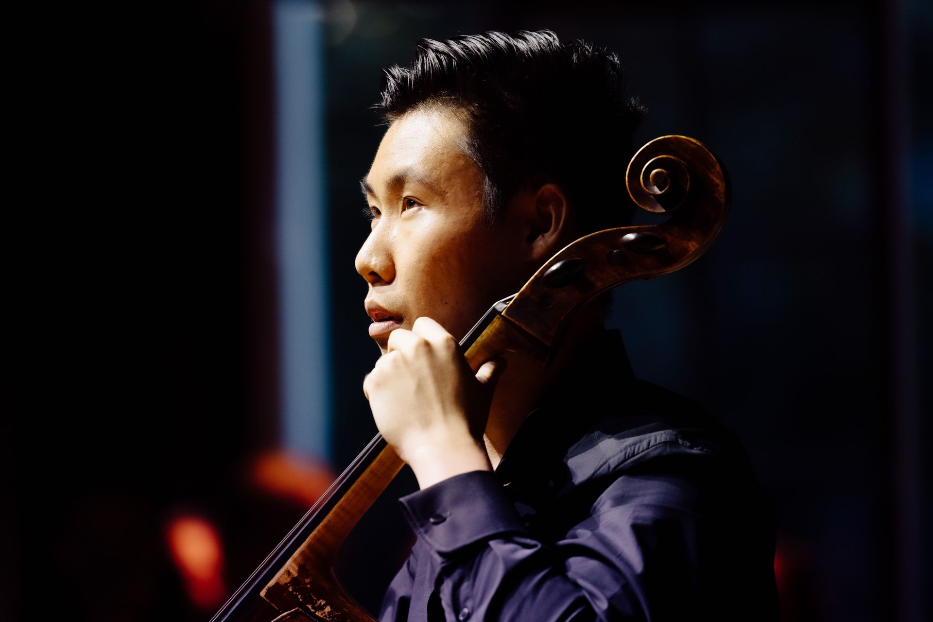 Michael Song holding his cello, lit from the side against a dark stage